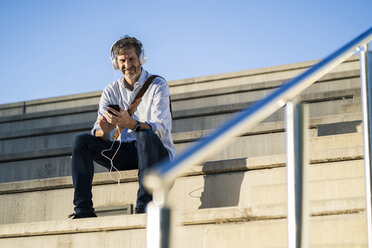 Porträt eines lächelnden reifen Mannes, der mit Kopfhörern und Mobiltelefon auf den Stufen sitzt - GIOF04919