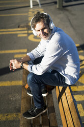 Mature man sitting on a bench listening to music with headphones - GIOF04928