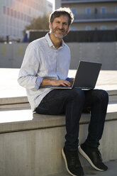 Portrait of smiling mature man sitting in the city using laptop - GIOF04933