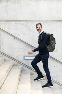 Lächelnder reifer Mann mit Rucksack beim Treppensteigen in der Stadt - GIOF04939
