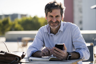 Portrait of smiling mature man sitting at outdoor table with cell phone and notebook - GIOF04950