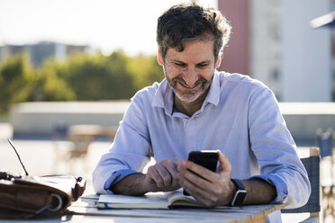 Lächelnder reifer Mann sitzt an einem Tisch im Freien mit Handy und Notizbuch - GIOF04951