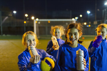 Porträt einer lächelnden Mädchenfußballmannschaft, die eine Trainingspause einlegt und nachts auf dem Spielfeld Wasser trinkt - HOXF04193