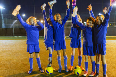 Porträt einer selbstbewussten Mädchenfußballmannschaft mit Wasserflaschen, die nachts auf dem Spielfeld jubeln - HOXF04198