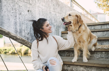 Smiling young woman stroking her Golden retriever dog on stairs outdoors - RAEF02233