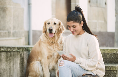 Young woman with her Golden retriever dog on stairs outdoors using cell phone - RAEF02239
