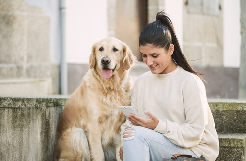 Junge Frau mit ihrem Golden Retriever Hund auf einer Treppe im Freien mit Handy - RAEF02239