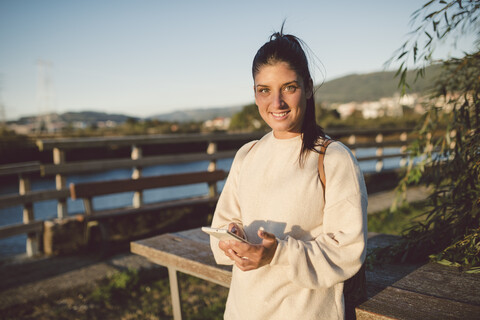 Portrait of smiling young woman with cell phone at the waterfront stock photo