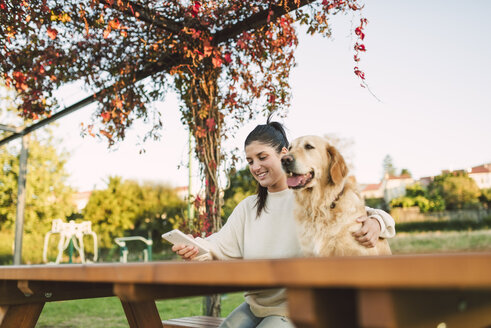 Lächelnde junge Frau mit Handy und ihrem Golden Retriever Hund, die sich in einem Park ausruhen - RAEF02253