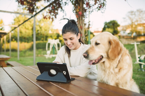 Lächelnde junge Frau, die in einem Park mit ihrem Hund ein Tablet benutzt, lizenzfreies Stockfoto