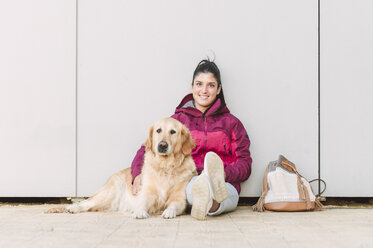 Porträt einer lächelnden jungen Frau mit ihrem Golden-Retriever-Hund, der an einer Wand sitzt - RAEF02261