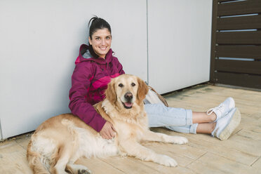 Portrait of a smiling young woman with her Golden retriever dog sitting at a wall - RAEF02263