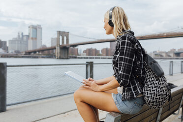 USA, New York City, Brooklyn, junge Frau sitzt mit Rucksack, Kopfhörern und Tablet am Wasser - BOYF01121