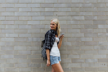 Lächelnde junge Frau mit Mobiltelefon an einer Backsteinmauer - BOYF01125