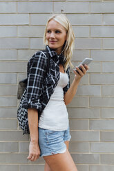 Lächelnde junge Frau mit Mobiltelefon an einer Backsteinmauer - BOYF01126