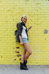 Porträt einer jungen Frau mit Handy an einer gelben Backsteinmauer - BOYF01130