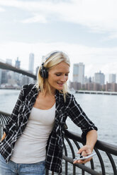 USA, New York City, Brooklyn, smiling young woman standing at the waterfront with headphones and cell phone - BOYF01147