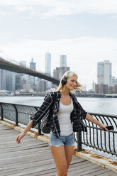 USA, New York City, Brooklyn, glückliche junge Frau mit Kopfhörern und Handy beim Tanzen am Wasser - BOYF01149