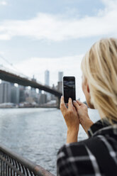 USA, New York City, Brooklyn, junge Frau am Wasser, die mit ihrem Handy ein Foto von der Brooklyn Bridge macht - BOYF01152