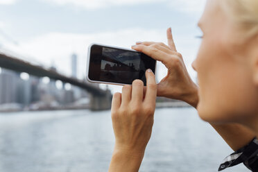 USA, New York City, Brooklyn, junge Frau am Wasser, die mit ihrem Handy ein Foto von der Brooklyn Bridge macht - BOYF01153