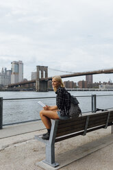 USA, New York City, Brooklyn, lächelnde junge Frau, die mit Rucksack und Tablet am Wasser sitzt - BOYF01159