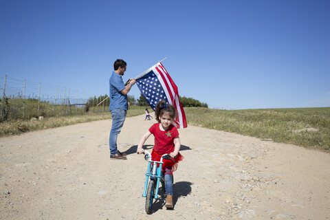 Mädchen mit Fahrrad und Mann mit amerikanischer Flagge auf Weg in abgelegener Landschaft, lizenzfreies Stockfoto