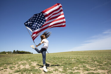 Happy girl holding American flag jumping on field in remote landscape - ERRF00196