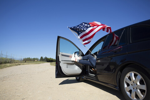 Mädchen mit amerikanischer Flagge im Auto sitzend auf einem Weg in abgelegener Landschaft, lizenzfreies Stockfoto