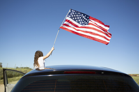 Mädchen hält amerikanische Flagge aus einem Auto unter blauem Himmel, lizenzfreies Stockfoto