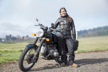 Porträt einer Motorradfahrerin in voller Länge, die eine Lederjacke trägt und neben einem Motorrad auf einem Feldweg steht - CAVF57472