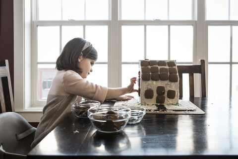 Seitenansicht eines Mädchens, das zu Hause ein Lebkuchenhaus auf einem Tisch am Fenster baut, lizenzfreies Stockfoto