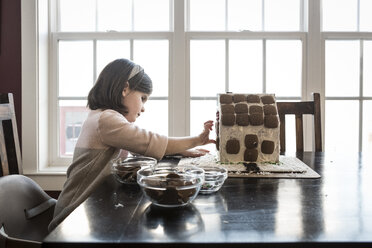 Seitenansicht eines Mädchens, das zu Hause ein Lebkuchenhaus auf einem Tisch am Fenster baut - CAVF57537