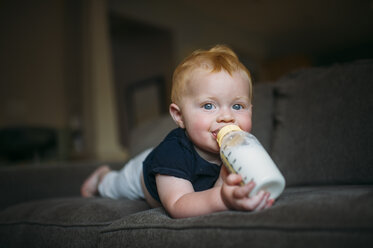 Porträt eines süßen kleinen Jungen, der Milch aus der Flasche trinkt, während er zu Hause auf dem Sofa liegt - CAVF57547