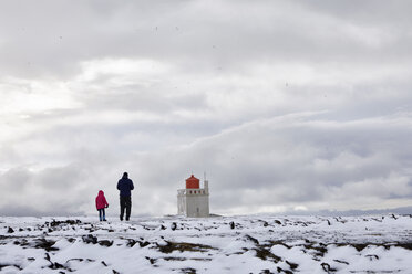 Rückansicht von Vater und Tochter, die auf einer verschneiten Landschaft vor einem Leuchtturm und einem bewölkten Himmel stehen - CAVF57554