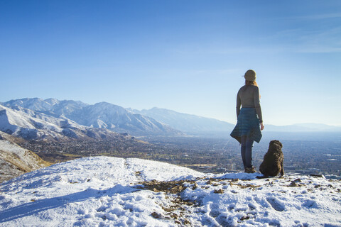 Rear view of woman with dog standing on mountain against sky during winter stock photo