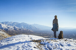 Rear view of woman with dog standing on mountain against sky during winter - CAVF57564
