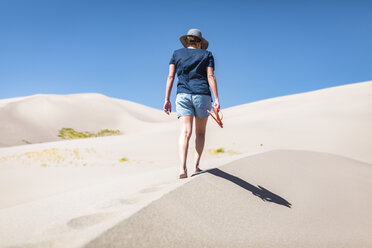 Rückansicht einer Frau, die einen Flip-Flop hält, während sie an einem sonnigen Tag im Great Sand Dunes National Park über den Sand läuft - CAVF57569