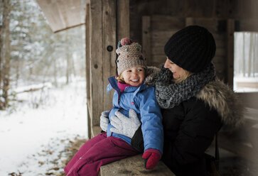 Happy mother standing by daughter sitting on window during winter - CAVF57606