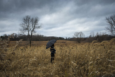 Teenager-Mädchen mit Regenschirm auf einem Feld vor stürmischen Wolken stehend - CAVF57607