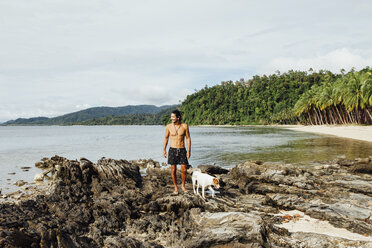 Hemdloser Wanderer mit Hund steht auf Felsen gegen den Himmel am Strand - CAVF57659