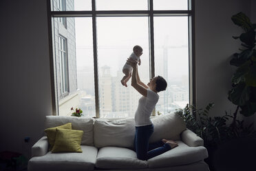 Seitenansicht einer glücklichen Mutter, die ihren Sohn hochhebt, während sie zu Hause auf dem Sofa am Fenster kniet - CAVF57698