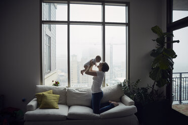 Seitenansicht einer glücklichen Mutter, die mit ihrem Sohn spielt, während sie zu Hause auf dem Sofa am Fenster kniet - CAVF57699