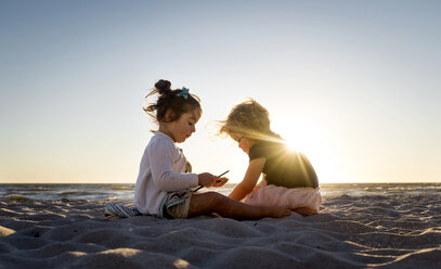 Schwestern sitzen auf Sand am Strand gegen klaren Himmel bei Sonnenuntergang - CAVF57717