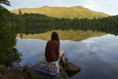 Rückansicht einer Frau, die auf einem Felsen an einem Teich im Wald sitzt und die Aussicht betrachtet, lizenzfreies Stockfoto