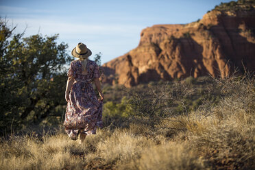Rückansicht einer Frau mit Hut, die auf einer Wiese vor dem Himmel spazieren geht - CAVF57781