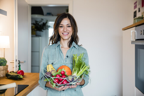 Frau steht in der Küche und hält einen Korb mit frischem Obst und Gemüse in der Hand, lizenzfreies Stockfoto