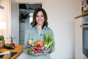 Frau steht in der Küche und hält einen Korb mit frischem Obst und Gemüse in der Hand - MOEF01711