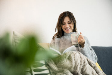 Woman sitting on couch, wrapped in a blanket, reading book, drinking tea - MOEF01749