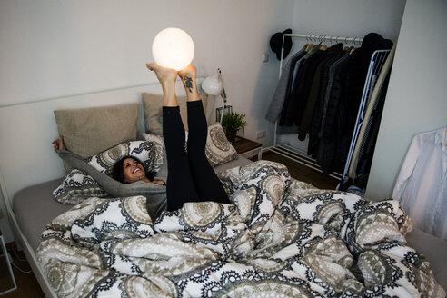 Frau liegt auf dem Bett und balanciert eine kugelförmige Lampe auf ihren Füßen - MOEF01752