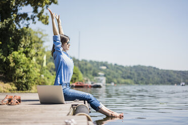 Frau mit Laptop auf einem Steg an einem See sitzend, sich streckend - MOEF01770